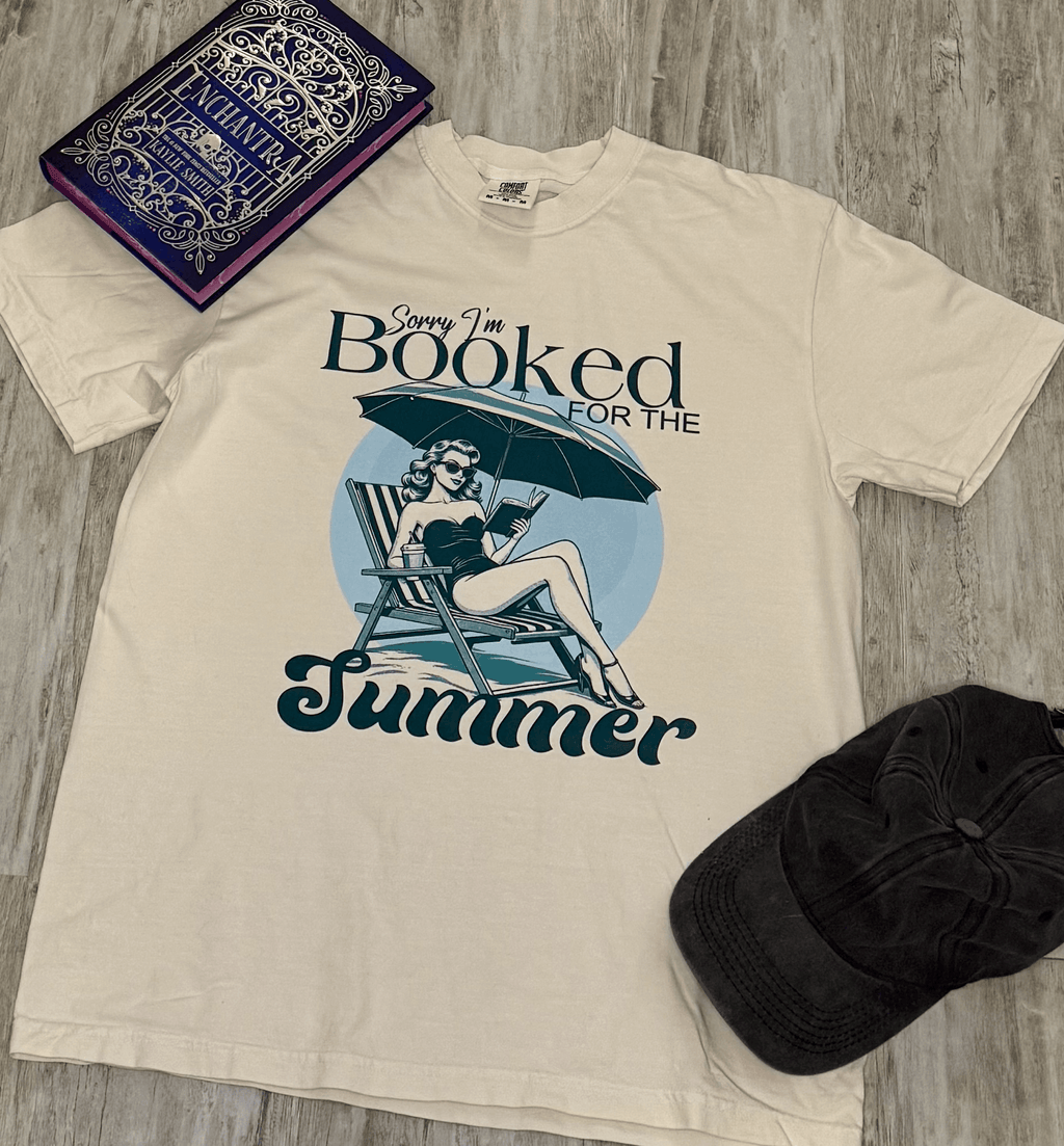 Beige t-shirt with 'Sorry I'm Booked for the Summer' graphic on a wooden surface with a book and black cap.