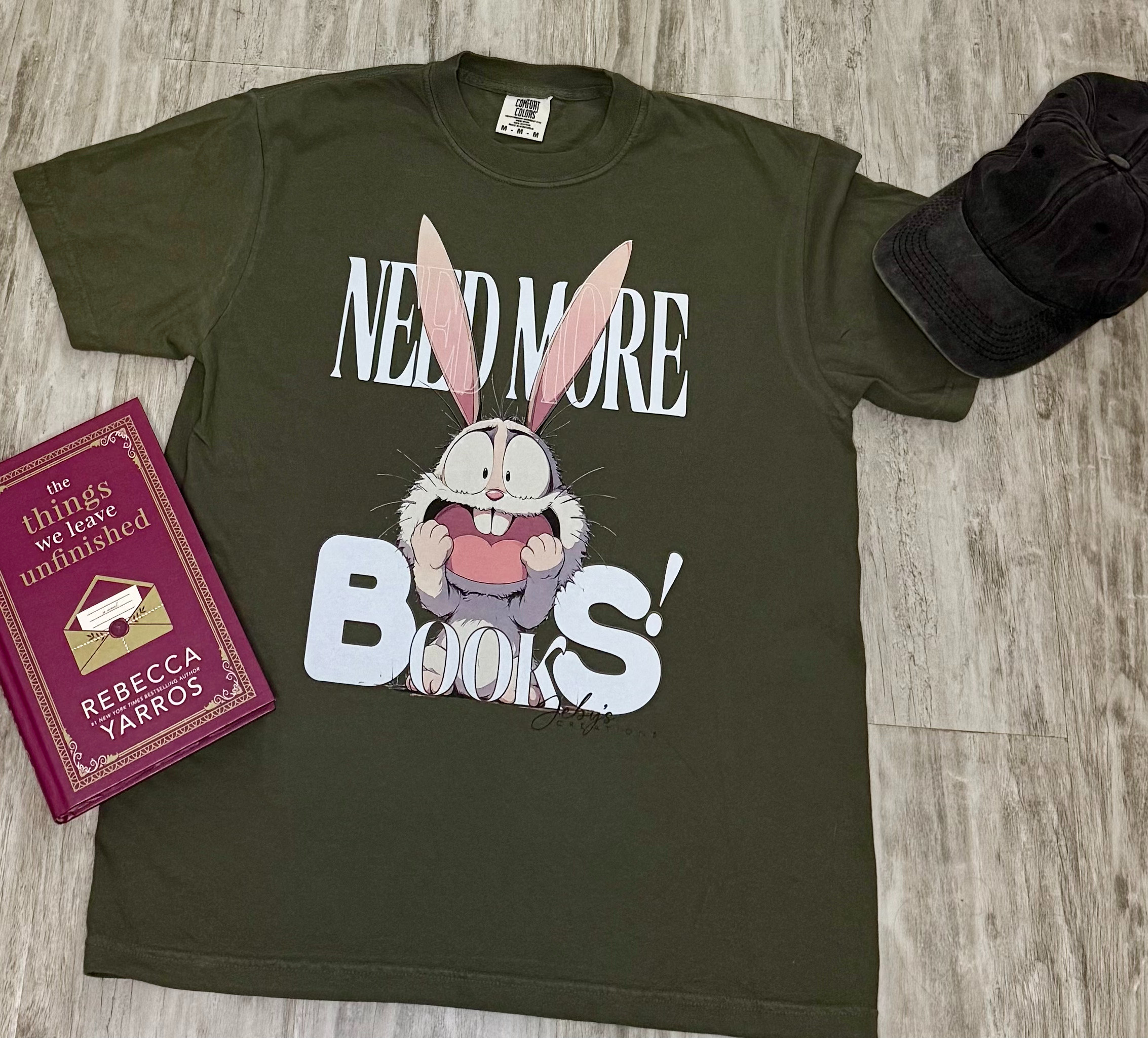 Green t-shirt with cartoon character and 'Need More Books!' text on a wooden surface with a book and cap.