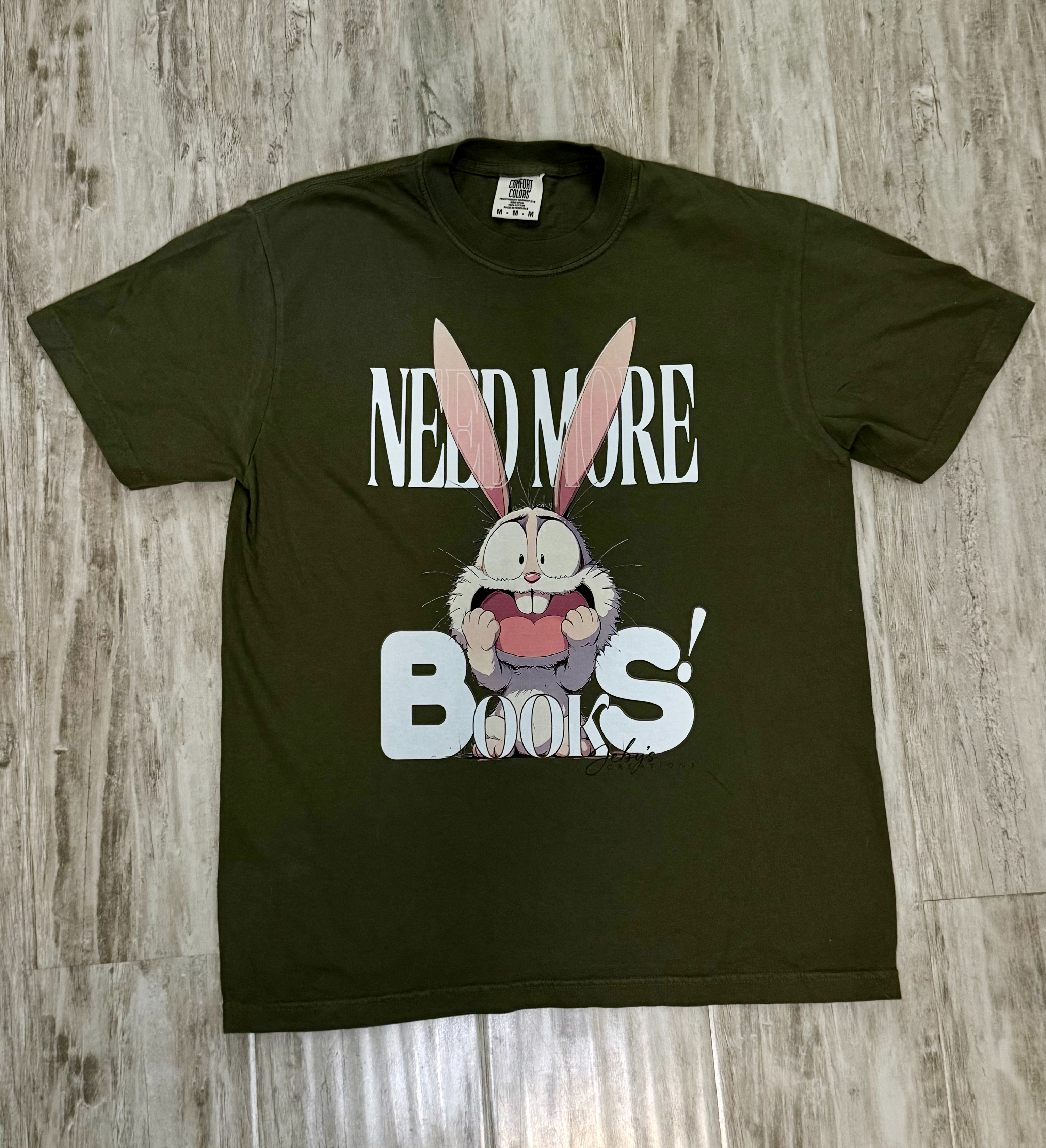 Green t-shirt with cartoon character and text 'NEED MORE BOOKS!' on a wooden surface