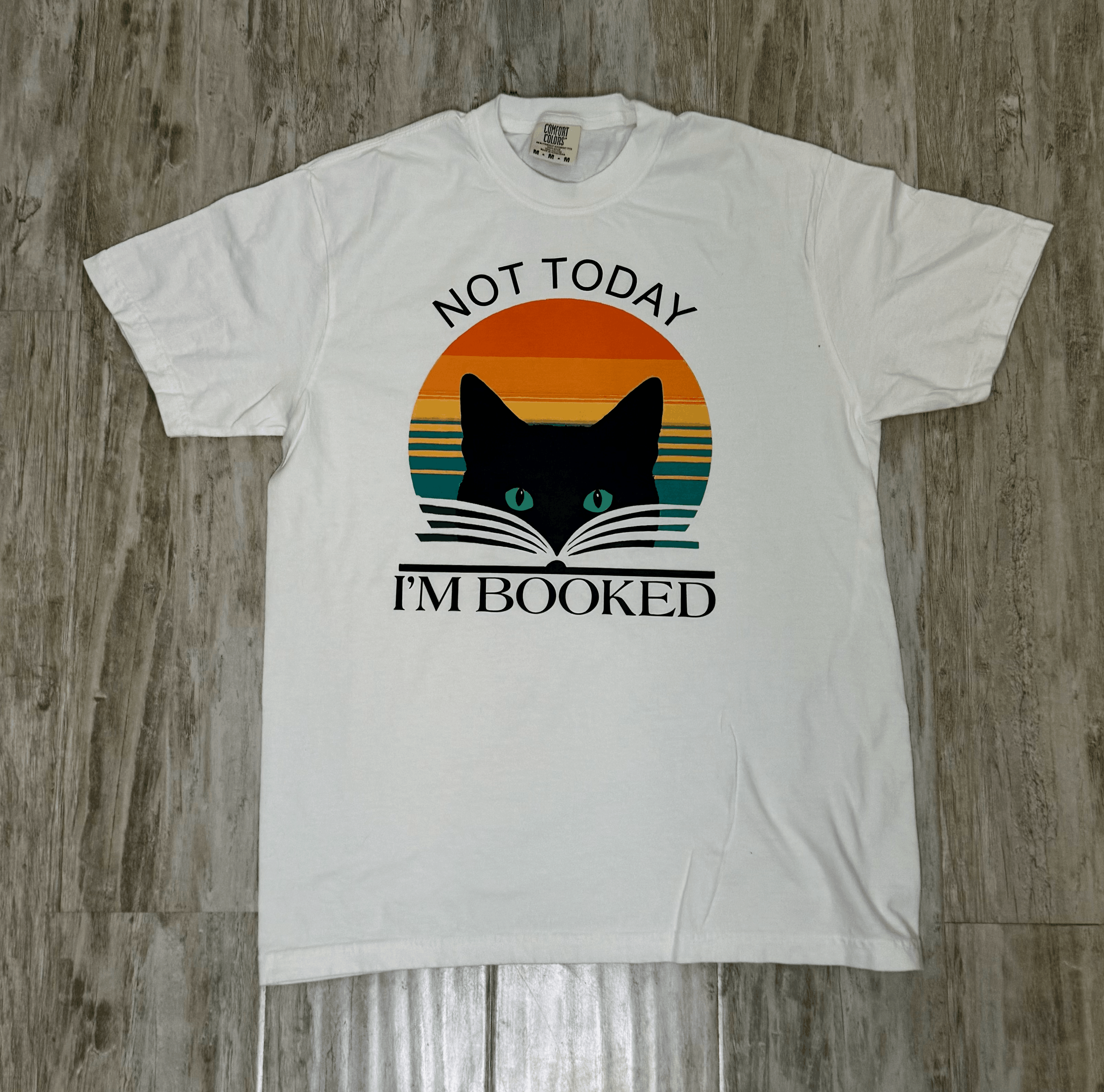White t-shirt with a cat and book graphic and text 'Not Today, I'm Booked' on a wooden surface.