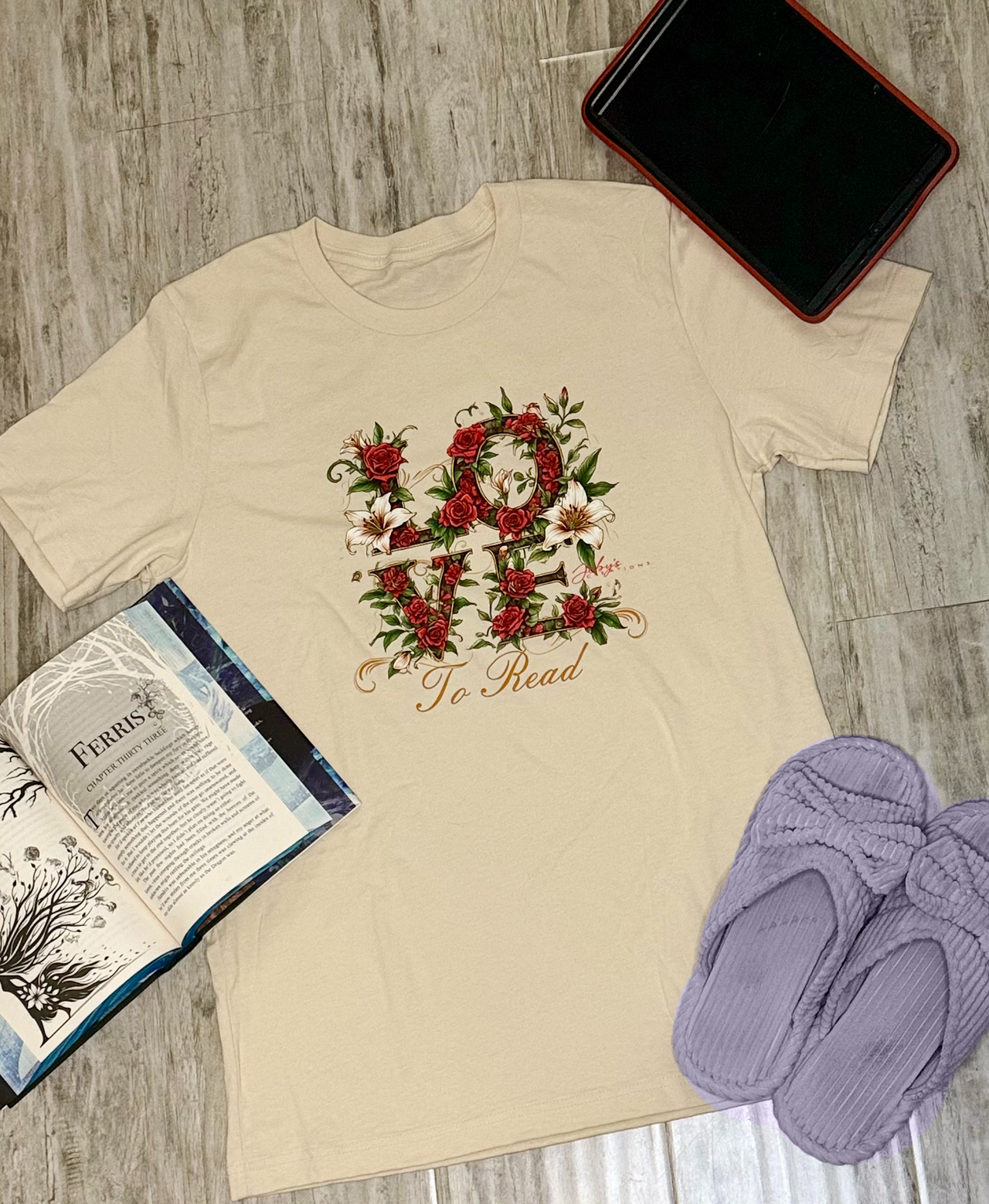 Beige t-shirt with floral design and text on a wooden surface with a phone, book, and slippers.
